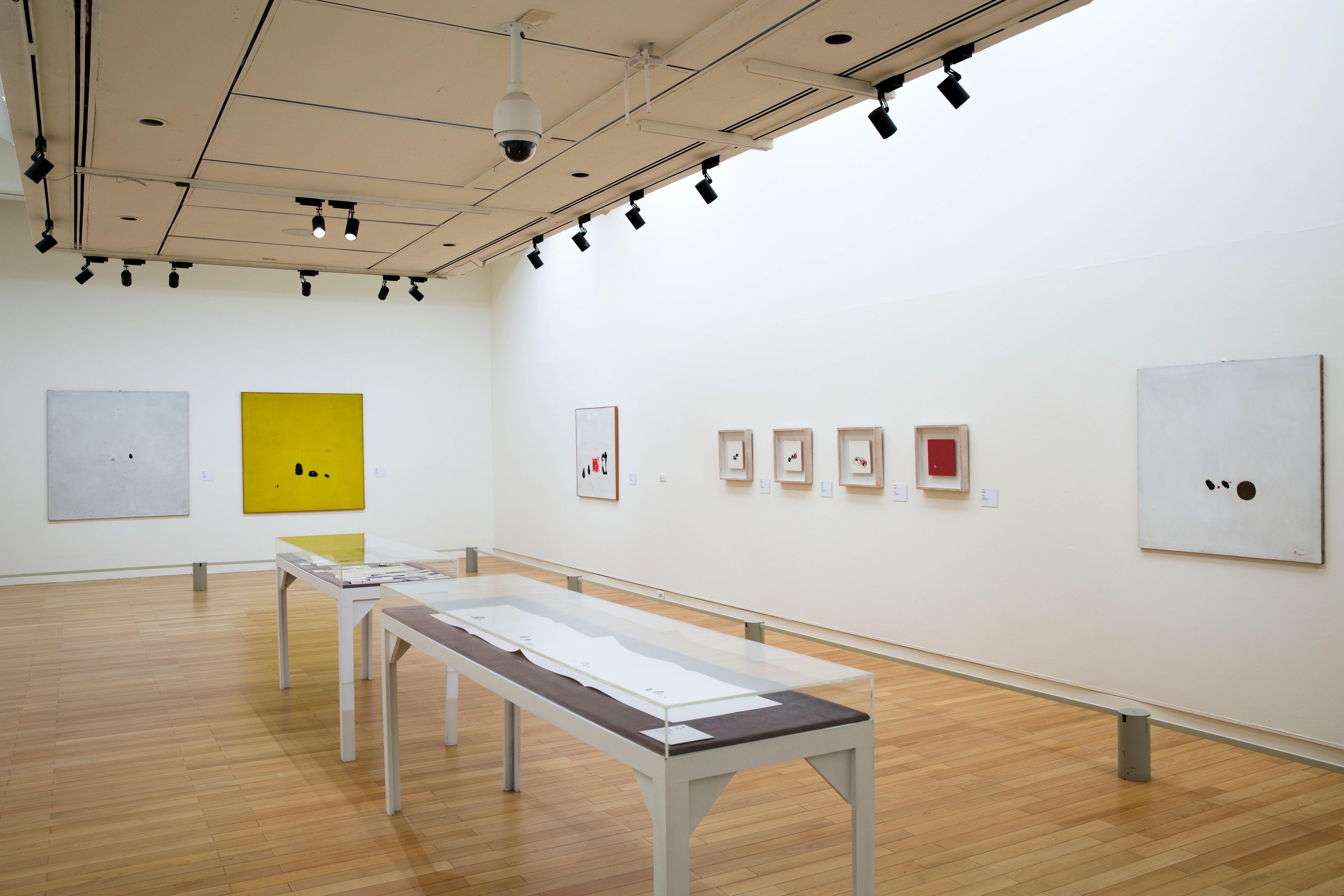 Installation. Two long cases with works on paper are in the middle of a gallery hung with works of art. Most of the compositions are square and dominated by white, yellow, or red, which act as backgrounds for small spots and marks in black or other colours.