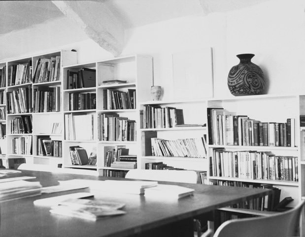 Black and white photograph. Book-lined shelves fill the wall beyond a conference-sized study desk.
