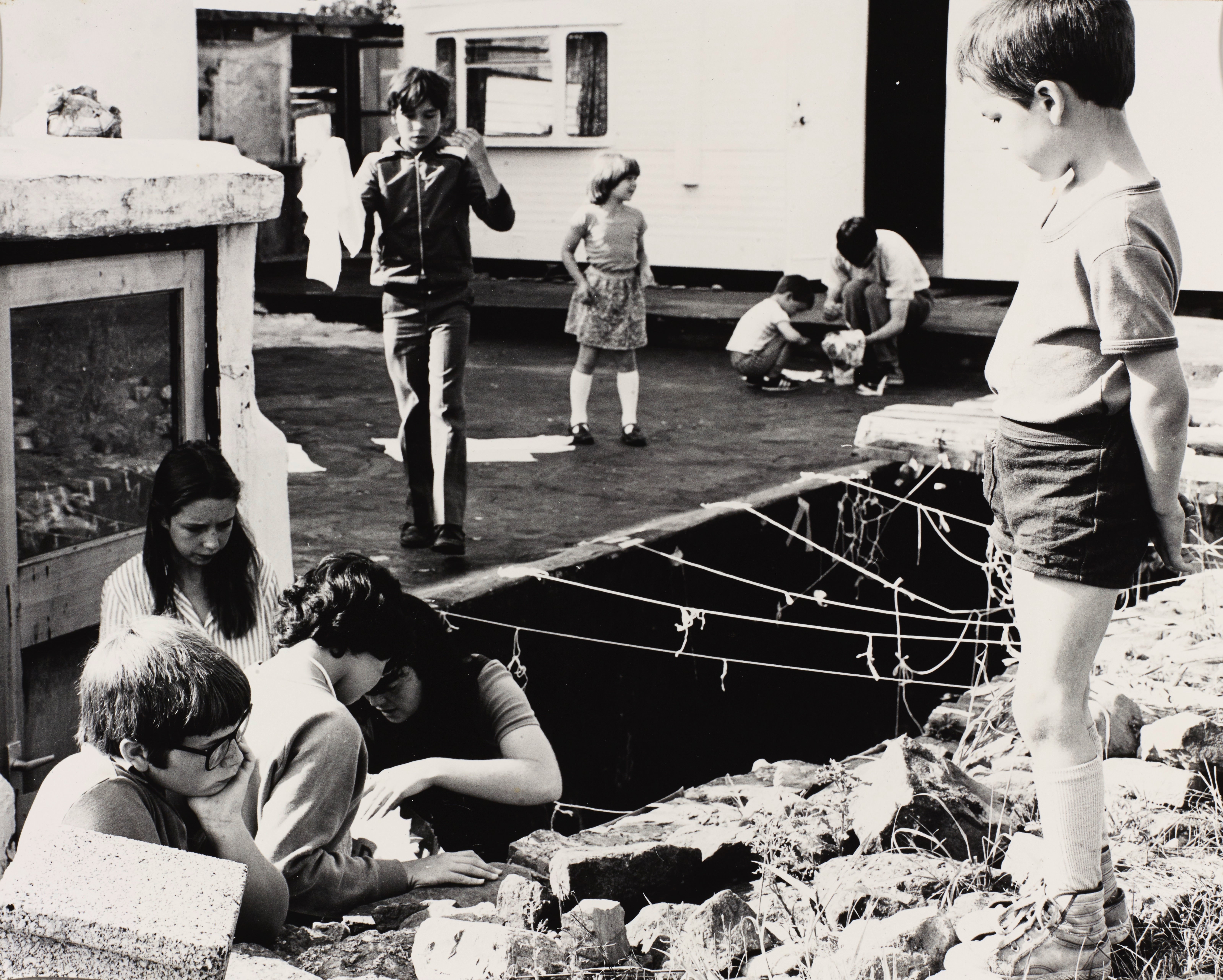 Black and white photograph. Nine children are around or in a trench in a courtyard.