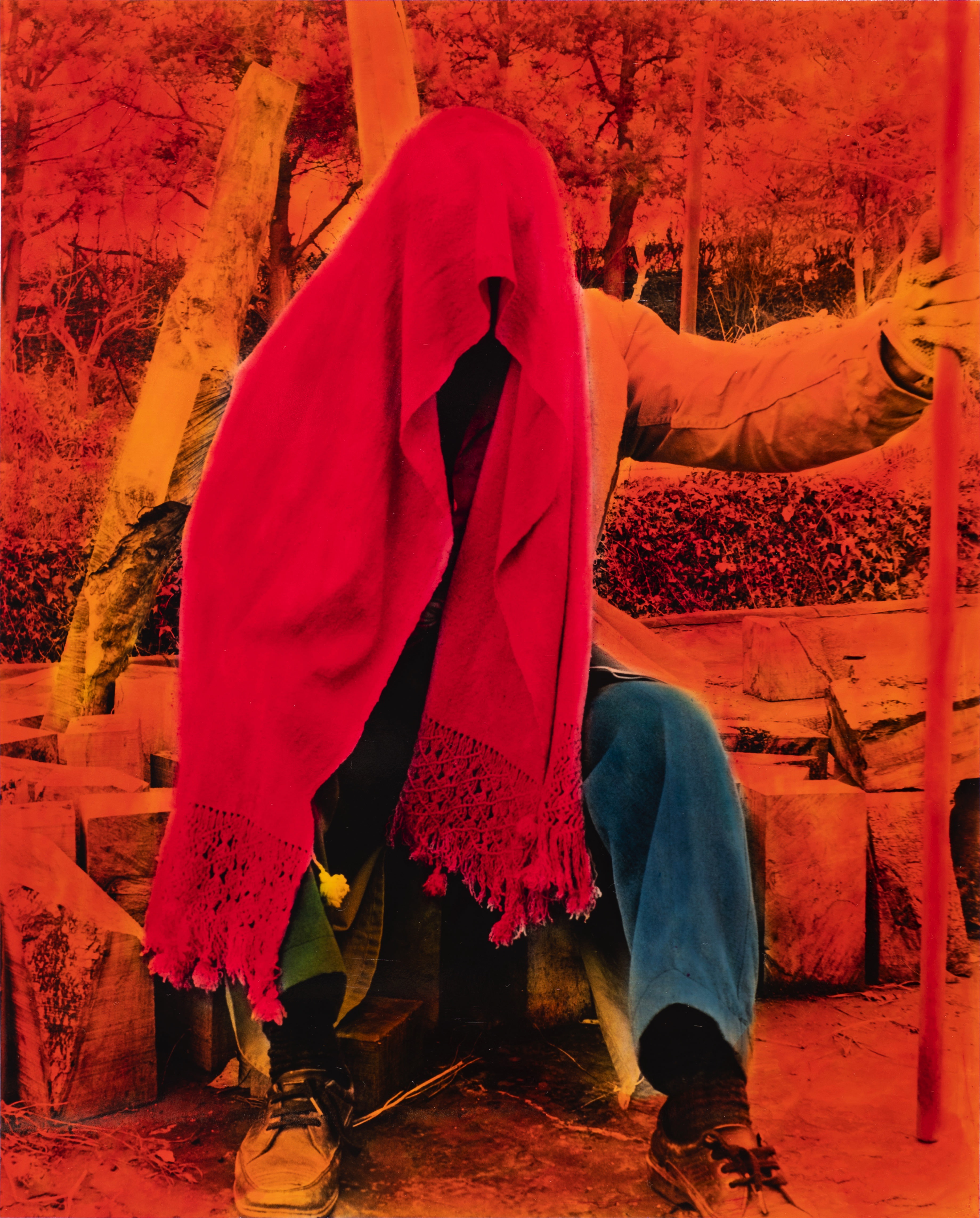 Hand-coloured black and white photograph. A person sits with a scarlet-red scarf draped over their head, obscuring their face. One hand is out and holds a staff. The background is painted traffic-cone orange and the pants indigo blue.