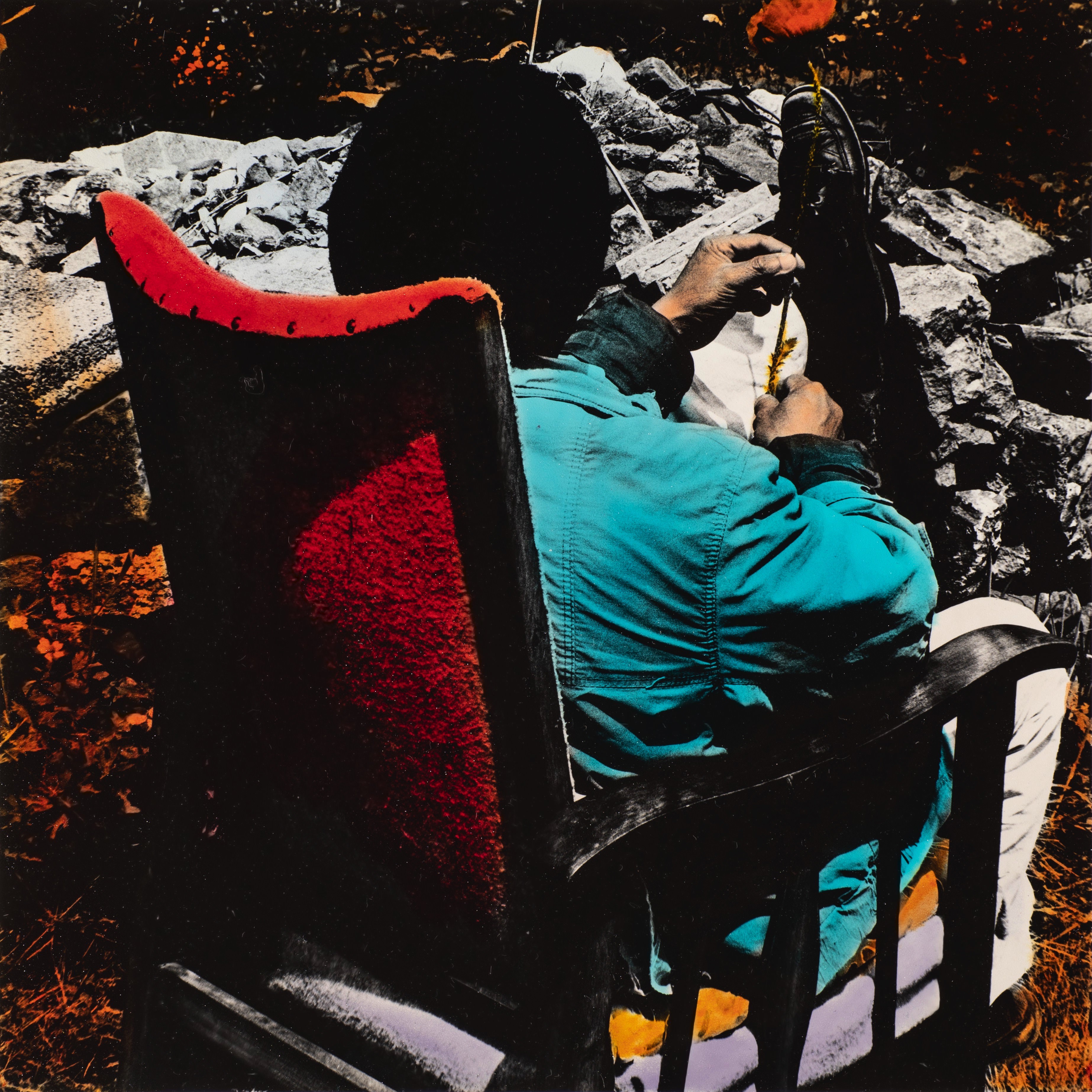 Hand-coloured black and white photograph. A man sits facing away from us in a high-backed armchair. The back of the chair is vivid red, and the jacket he wears is turquoise. One foot is on the ground and the other foot is propped up on a rock. The person holds something slender, perhaps a twig, in his peach-toned hands.