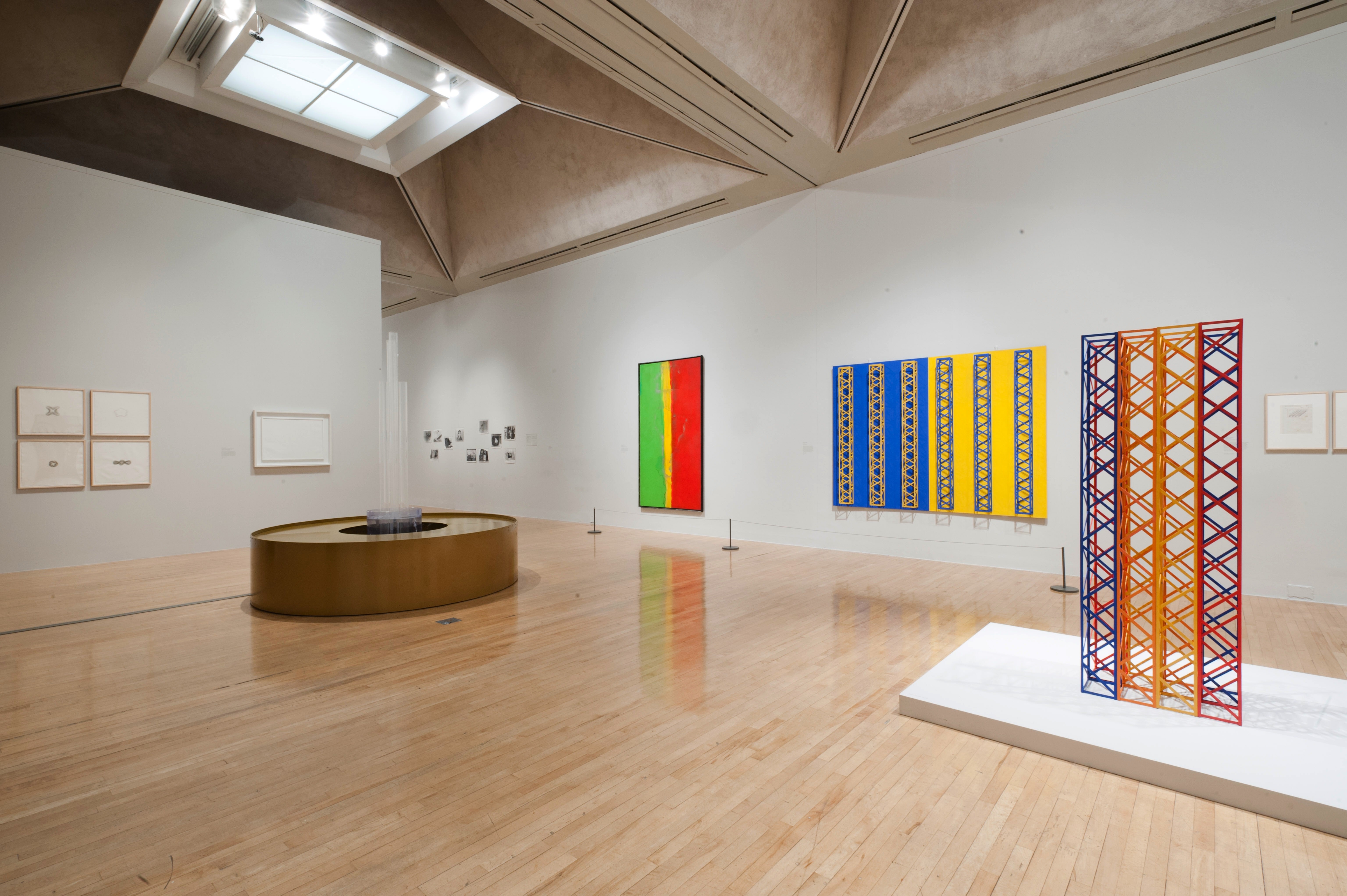 Installation. Two sculptures are in the centre of a gallery hung with mostly colourful works of art, many of which create patterns. The gallery itself has shiny wood floors and trapezoid-shaped coffers set into the ceiling.