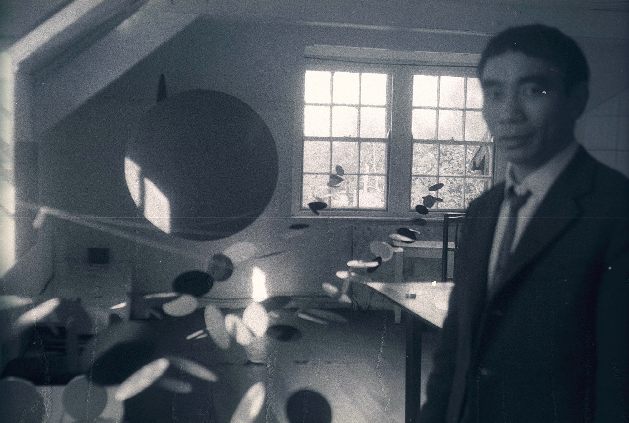 Black and white photograph. A person, Li Yuan-chia, stands close to us in his studio. He is blurry because the camera’s focus is on the end of a long path of fluttering discs that lead back to windows on the far side of the room.