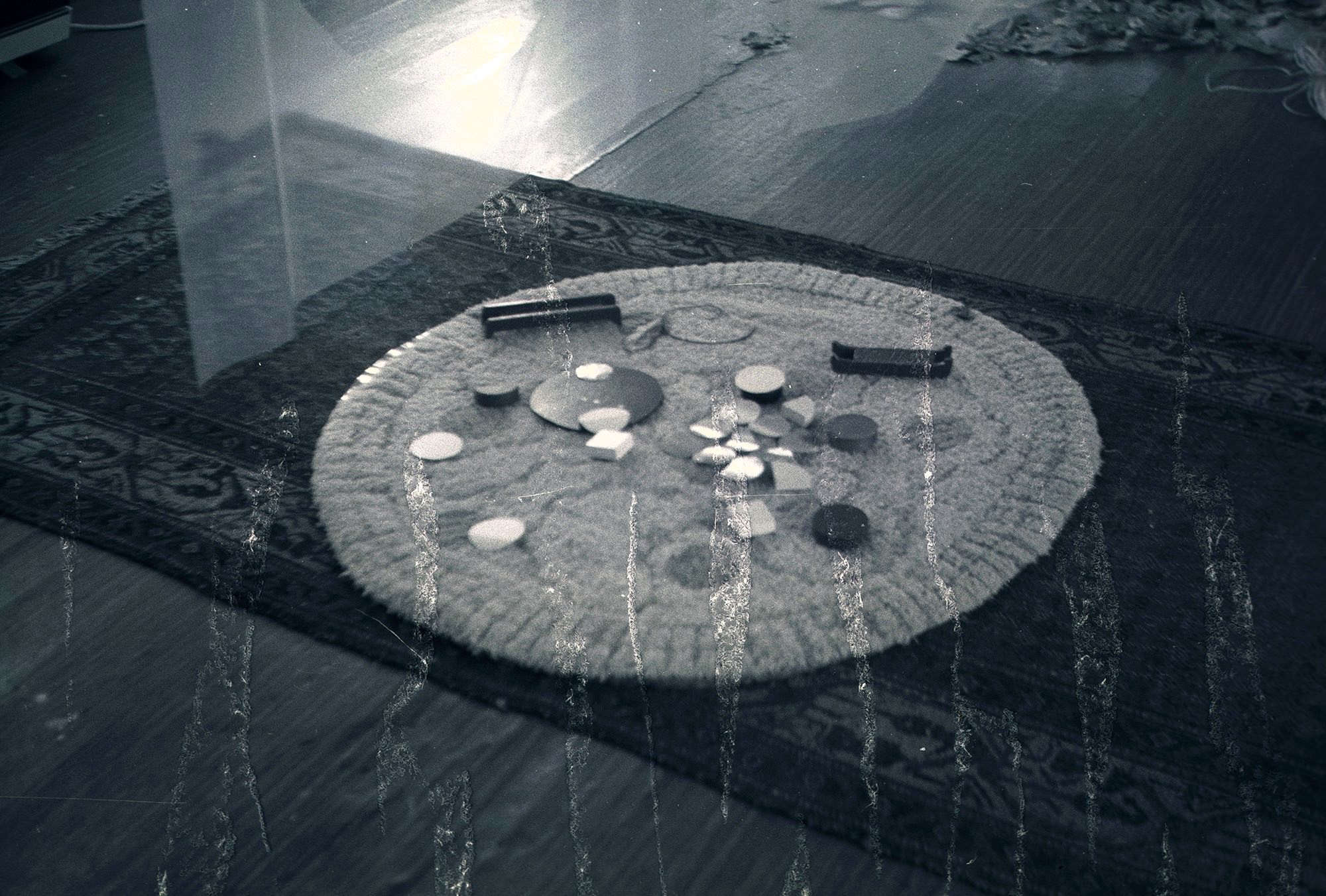 Black and white photograph. Discs are laid out on a round area rug placed on top of a rectangular area rug. Texture on the surface of the photograph flickers like flames across the bottom of the image.