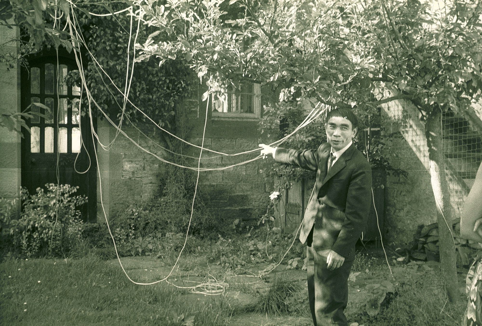 Black and white photograph. A man wearing a suit smiles at someone to our right as he points to strings hanging from trees in an overgrown courtyard. He has a bowl haircut, thick brows, and dimples.