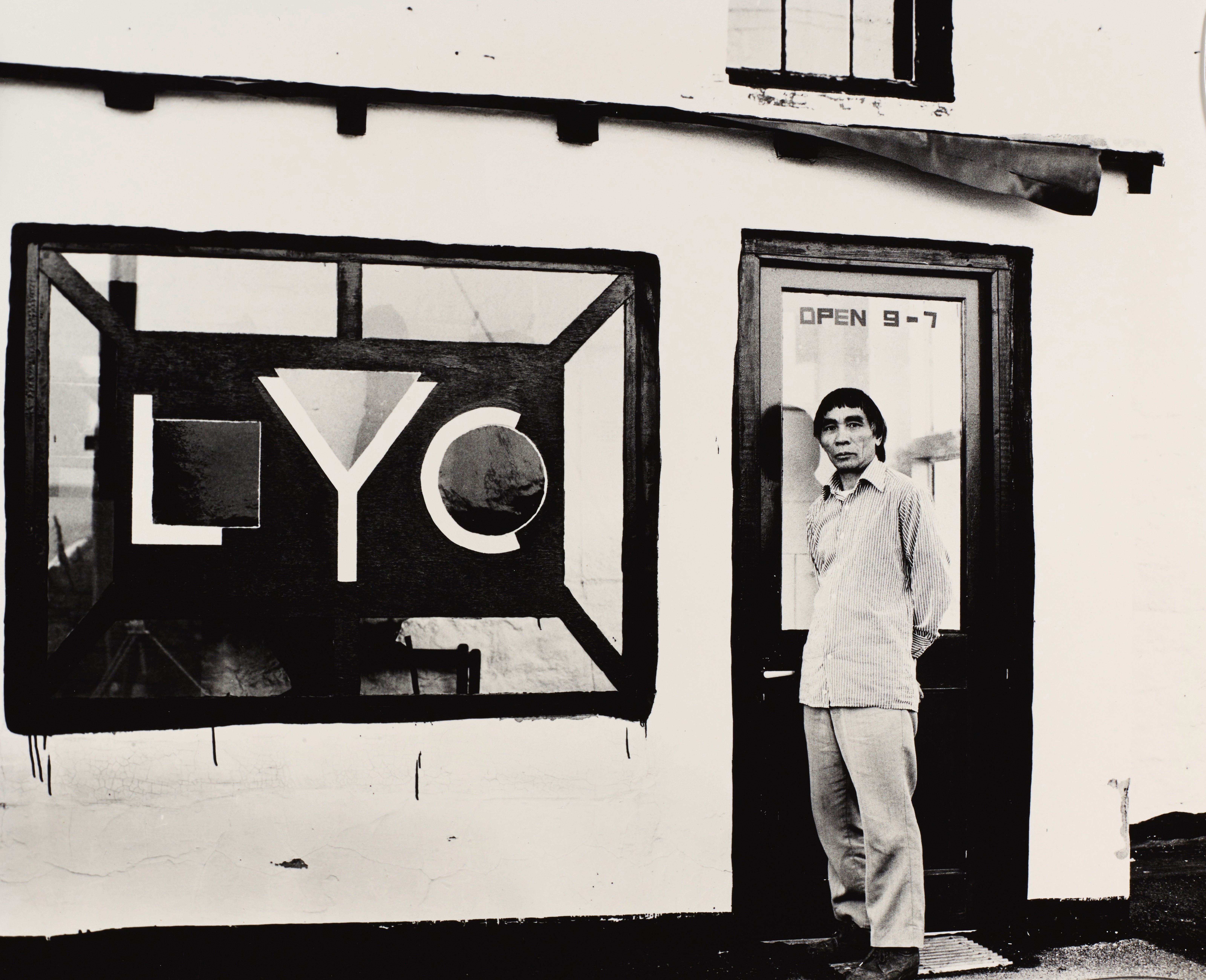 Black and white photograph. A man stands in front of a door, hands behind his back, looking at us. Text applied to the glass in the top half of the door reads “Open 9-7”. The storefront window to our left has the initials “LYC” in a geometric design. The L nestles a square in its bend, the Y a triangle, and the C a circle.