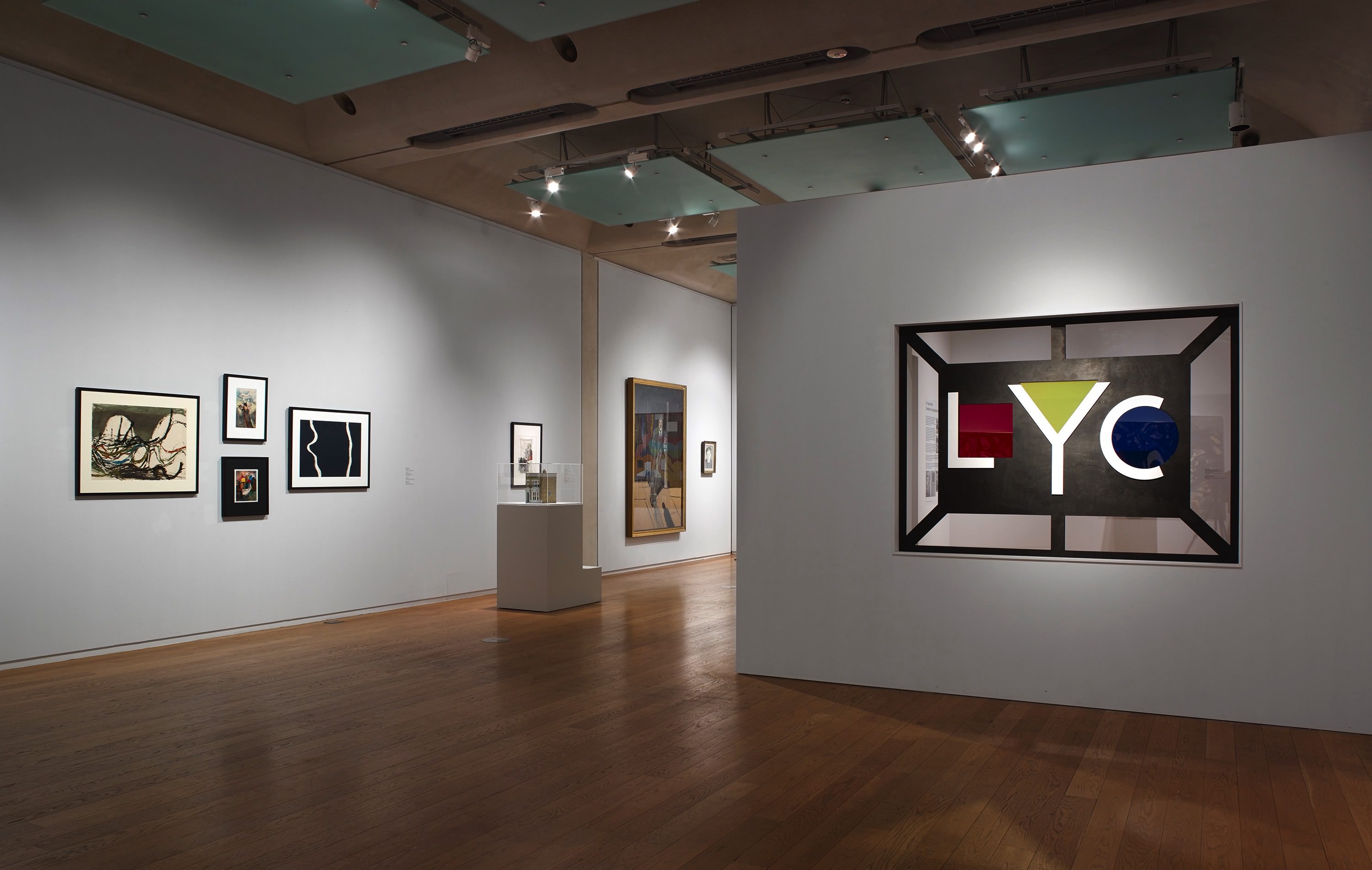 Installation. Five works of art on two walls in the space we occupy are spotlighted in a dimmed gallery. A sign across from us shows geometric shapes tucked into initials: a red square in a capital L, a yellow triangle filling the top of a Y, and a blue circle hugged by a C.